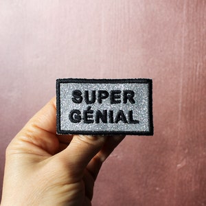 May include: A silver glitter patch with black embroidered text that reads "SUPER GÉNIAL".