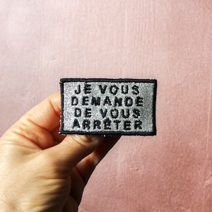 May include: A silver and black embroidered patch with the text "Je vous demande de vous arreter" in black.