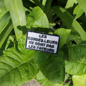 Op de afbeelding: Een witte patch met zwarte tekst die "Les Conseillers Ne Sont Pas Les Payeurs" luidt, is geplaatst op een groene, bladerrijke plant.