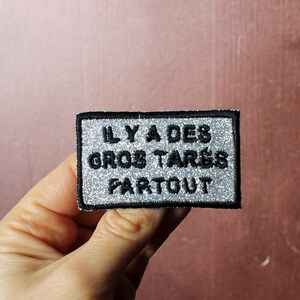 May include: A silver glitter iron-on patch with black text that reads "IL Y A DES GROS TARES PARTOUT".