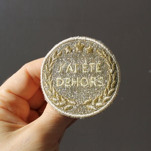 Może przedstawiać: Okrągła naszywka z haftem z francuskim tekstem "J'AI ÉTÉ DEHORS" złotą nicią. Naszywka ma srebrne, brokatowe tło, złoty wieniec laurowy i trzy złote gwiazdy u góry.