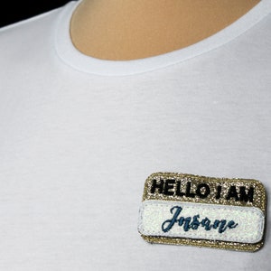 Puede incluir: Camiseta blanca con una etiqueta con el texto "HELLO I AM Insane". La etiqueta tiene un borde dorado con purpurina y un centro blanco con letras azules. La camiseta es lisa.