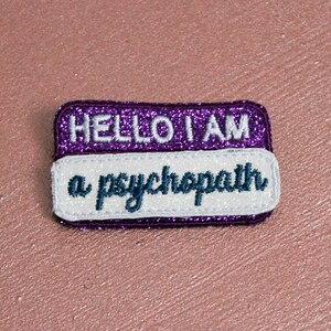 May include: A rectangular name tag patch with the text "HELLO I AM" above "a psychopath". The patch has a purple glitter border and a white section with teal lettering. The background is a pinkish-brown color.