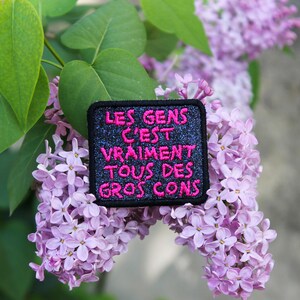 Peut inclure: Un écusson noir avec du texte rose qui dit "LES GENS C'EST VRAIMENT TOUS DES GROS CONS" est présenté sur un fond de fleurs de lilas violettes et de feuilles vertes. L'écusson a une texture scintillante.