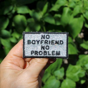 Könnte beinhalten: Ein rechteckiger Aufnäher mit dem Text "NO BOYFRIEND NO PROBLEM" in schwarzen Großbuchstaben. Der Aufnäher hat einen silbernen Glitzerhintergrund und einen schwarzen Rand. Der Aufnäher wird vor einem Hintergrund aus grünen Blättern gehalten.