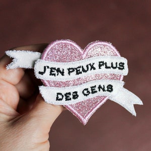 Peut inclure: Un écusson en forme de cœur rose pailleté avec le texte français "J'EN PEUX PLUS DES GENS" brodé sur une bannière blanche. L'écusson est tenu dans une main sur un fond marron.