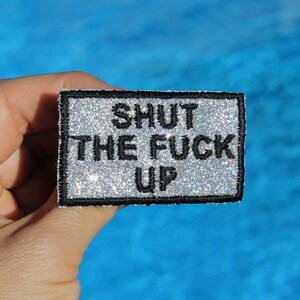 May include: A rectangular patch with a silver glitter background and black border. The patch features the phrase "SHUT THE FUCK UP" in bold, black capital letters. The patch is held against a blue background.