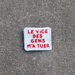 May include: A white patch with red embroidered text that reads "LE VICE DES GENS M'A TUER". The patch has a white border and is set against a textured gray surface. The text is in a bold, sans-serif font.