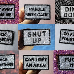 Peut inclure: Un ensemble de neuf écussons rectangulaires thermocollants avec un fond à paillettes argentées et du texte brodé en noir. Les phrases incluent STAY AWAY FROM ME, HANDLE WITH CARE, DING DONG, PUCK EVERYBODY, SHUT UP, GO FUCK YOURSELF, FUCK EVERYTHING, CAN I GET AN AMEN et FUCK IT.