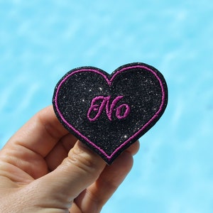 May include: A black glitter heart-shaped patch with a pink border and the word "No" embroidered in pink. The patch is held against a light blue background.
