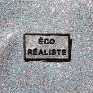 May include: A rectangular embroidered patch with a silver glitter background and a black border. The patch features the black embroidered text ÉCO RÉALISTE centered in two lines. The design is clean and features a high-contrast color scheme.