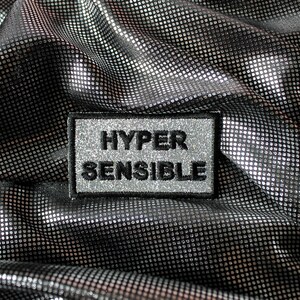 May include: A silver and black fabric with a repeating dot pattern. A rectangular patch with the words "HYPER SENSIBLE" in black lettering is sewn onto the fabric. The patch has a silver glitter border.