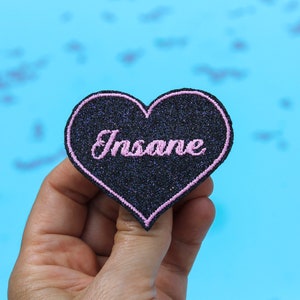 Puede incluir: Un parche negro en forma de corazón con la palabra "Insane" bordada en rosa. El parche tiene un borde rosa y una textura brillante. El fondo es de color azul claro sólido.