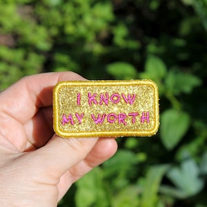 Puede incluir: Un parche rectangular dorado con purpurina con la frase bordada "I KNOW MY WORTH" en letras rosas. El parche tiene un borde dorado y se sostiene sobre un fondo verde borroso.
