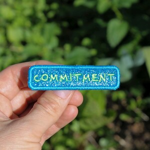May include: A rectangular, turquoise patch with the word "COMMITMENT" embroidered in green. The patch has a glittery border and is held against a blurred green background.