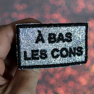 Peut inclure: Un patch en paillettes argentées avec du texte brodé en noir qui dit "A BAS LES CONS".