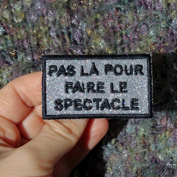 NOT THERE TO MAKE A SHOW brooch in silver sequined fabric with rainbow highlights, embroidered message in French, black thread, made in France