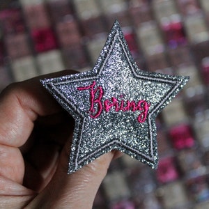 May include: A silver glitter star-shaped patch with a black border and the word "Boring" embroidered in pink. The patch is held up against a blurred background of various items.