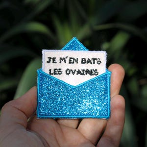 Può includere: Un cerotto a forma di busta blu glitterata con un inserto di carta bianca che dice "Je m'en bats les ovaires".