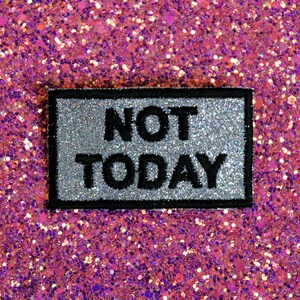 Puede incluir: Un parche de plancha de hierro plateado y negro con el texto "NOT TODAY" sobre un fondo de purpurina rosa.