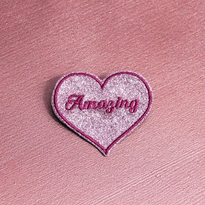Puede incluir: Un parche en forma de corazón de purpurina rosa con la palabra "Amazing" bordada en rosa.