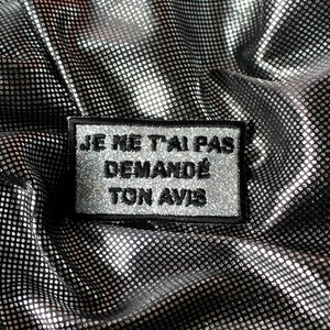 May include: A silver and black patch with the French text "JE NE T'AI PAS DEMANDÉ TON AVIS" embroidered in black and silver thread. The patch is rectangular with a black border and is set against a shiny, textured silver fabric.