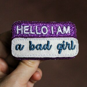 May include: A rectangular patch with the text "HELLO I AM" in white embroidered letters on a purple glitter background. Below, the text "a bad girl" is embroidered in teal on a white background.