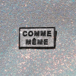 Peut inclure: Un écusson rectangulaire avec un fond à paillettes argentées et une bordure noire. Le texte COMME MÊME est brodé en lettres capitales noires épaisses au centre de l'écusson, posé sur une surface à paillettes argentées assortie.
