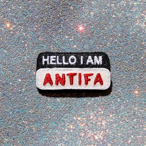 Könnte beinhalten: Ein rechteckiger gestickter Aufnäher mit einem schwarzen oberen Bereich mit dem weißen Text HELLO I AM und einem weißen unteren Bereich mit dem roten Text ANTIFA. Der Aufnäher hat abgerundete Ecken und einen gestickten Rand wie ein Namensschild.