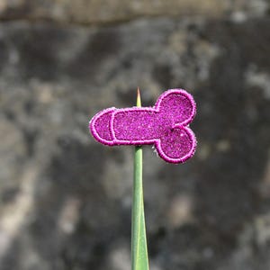 Puede incluir: Un aplique magenta brillante en forma de falo estilizado, montado sobre un tallo verde. El aplique tiene una superficie texturizada y brillante y un borde blanco. El fondo es borroso, de tono neutro.