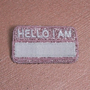 Puede incluir: Una etiqueta de nombre de purpurina rosa con el texto "HELLO I AM" y un espacio en blanco para escribir un nombre.
