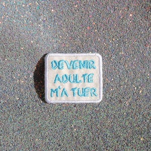 May include: A rectangular embroidered patch with a light beige background and a white border. The patch features the text DEVENIR ADULTE M'A TUER stitched in bright blue thread, arranged in three lines centered on the fabric surface.