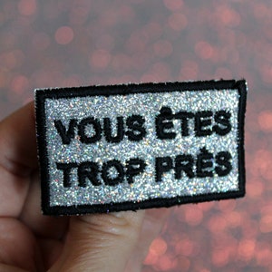Könnte beinhalten: Ein silberner Glitzer-Aufnäher mit schwarzem gesticktem Text, der "VOUS ÊTES TROP PRÈS" lautet.