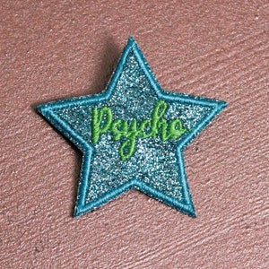 Könnte beinhalten: Ein teal-farbener Glitzer-Stern-Aufnäher mit teal-farbenem Rand und dem Wort "Psycho" in grüner Schreibschrift gestickt. Der Aufnäher befindet sich auf einer strukturierten, rötlich-braunen Oberfläche.