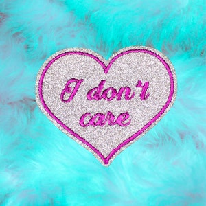 May include: A heart-shaped patch with a silver glitter finish and a bright pink embroidered border. The text "I don't care" is embroidered in bright pink cursive script across the center of the heart, which is set against a bright turquoise faux fur.