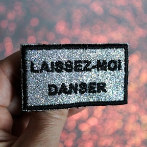 Könnte beinhalten: Ein silberner Glitzer-Aufnäher mit schwarzen Nähten und dem Text "LAISSEZ-MOI DANSER" in Schwarz.