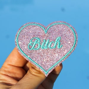 Może przedstawiać: Naszywka w kształcie serca z błyszczącą różową powierzchnią i turkusową obwódką. Słowo "Bitch" jest wyhaftowane turkusowym pismem pośrodku. Naszywka jest trzymana na jasnoniebieskim tle.