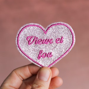 May include: A heart-shaped patch with a glittery silver surface and a bright pink border. The patch features the embroidered French text "Vieux et fou" in pink. The patch is held against a soft pink background.