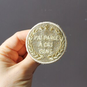 May include: Embroidered patch with the text "J'AI PARLÉ À DES CONS" in gold thread. The round patch has a laurel wreath border and a glittery appearance, suitable for clothing or accessories.