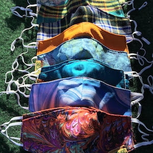 Puede incluir: Una pila de ocho mascarillas de tela coloridas con lazos el&aacute;sticos blancos para las orejas. Las mascarillas est&aacute;n hechas de diferentes patrones, incluyendo cuadros, flores y dise&ntilde;os abstractos. La mascarilla de abajo tiene el texto "www.SZMC" impreso en ella.