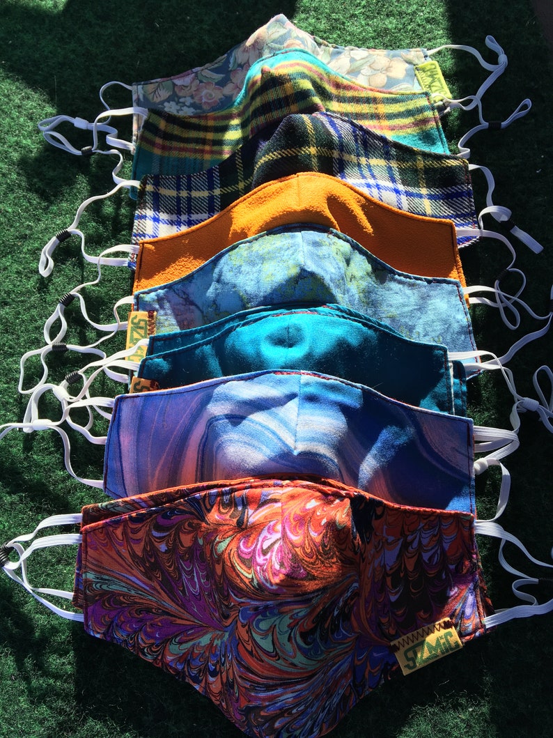 Puede incluir: Una pila de ocho mascarillas de tela coloridas con lazos el&aacute;sticos blancos para las orejas. Las mascarillas est&aacute;n hechas de diferentes patrones, incluyendo cuadros, flores y dise&ntilde;os abstractos. La mascarilla de abajo tiene el texto "www.SZMC" impreso en ella.