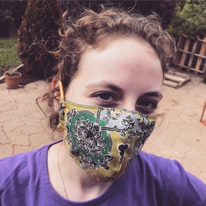 Puede incluir: Una persona usando una mascarilla facial con estampado amarillo, verde y negro. La mascarilla tiene un dise&ntilde;o floral.