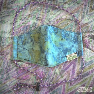 Puede incluir: Una mascarilla facial de patr&oacute;n azul y verde con una correa ajustable blanca y morada y una peque&ntilde;a etiqueta blanca con el texto "SZMIC" en ella.