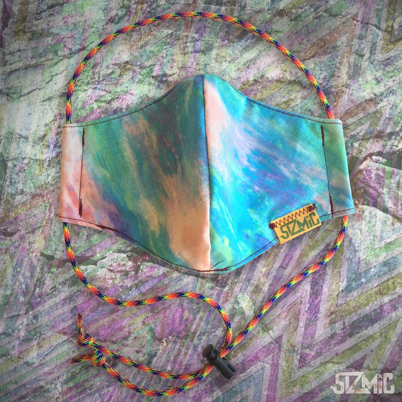 Puede incluir: Una mascarilla facial de tie-dye azul, verde y rosa con una correa ajustable marr&oacute;n y una peque&ntilde;a etiqueta marr&oacute;n que dice "SZMIC".