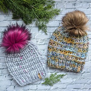 May include: Two handmade knit beanies with faux fur pom-poms. One is light gray with pink flecks and a magenta pom-pom. The other is multicolored with a brown pom-pom. Both have a small tag.