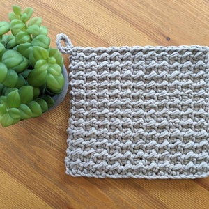 May include: A square, light grey crocheted potholder with a hanging loop. The potholder features a textured, woven design. A small green plant in a grey pot is to the left of the potholder on a wooden surface.