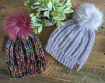 May include: Two knitted winter hats with faux fur pom-poms. One hat is a mix of rainbow colors, and the other is a light purple color. Both hats have a ribbed knit design and a small leather tag.