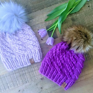 May include: Two cable-knit beanies with faux fur pom-poms. One is light purple with a light blue pom-pom, and the other is a vibrant purple with a brown pom-pom. Both have a small leather tag. The beanies are on a wooden surface.