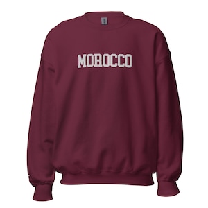 May include: A maroon sweatshirt with the word "MOROCCO" printed in white block letters across the chest. The sweatshirt has a crew neck and ribbed cuffs and hem. A tag is visible at the back of the neck.