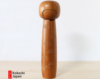 May include: A traditional Japanese Kokeshi doll carved from wood. The doll has a round head with painted eyes and a long, slender body with a carved floral pattern. The doll is made of natural wood and has a warm, rustic appearance.